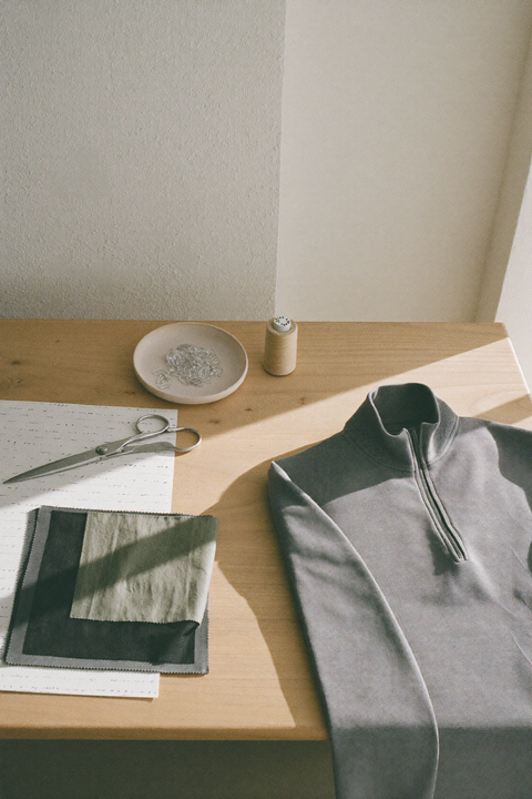 Ruhiger Ateliertisch aus Eiche mit einem ausgelegten Strick-Halfzip, einem unbeschrifteten Papierschnitt, einer Schneiderschere aus Stahl, einer Keramikschale mit Nadeln, Garn und Stoffmustern in Anthrazit und Salbei